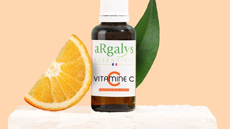 Une bouteille de vitamine C est posée sur un socle blanc, à côté d'une orange coupée en deux.